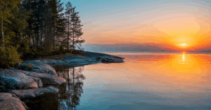 Picture taken from the shore of a mountain lake with trees in the foreground and the sun setting in the background.