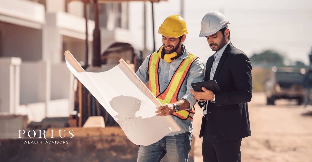 Two men wearing construction helmets outside a home under construction. Tey are looking at the plans for the building and seem to be discussing them. This is for the "selling a trades business in Lakeland FL" blog post. The Portus Wealth Advisors logo is in the bottom left of the image.
